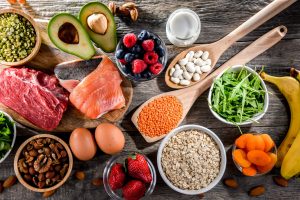 Assorted fertility diet foods including salmon, lean red meat, eggs, lentils, avocado, nuts, berries, oats, leafy greens, and bananas, displayed on a wooden table