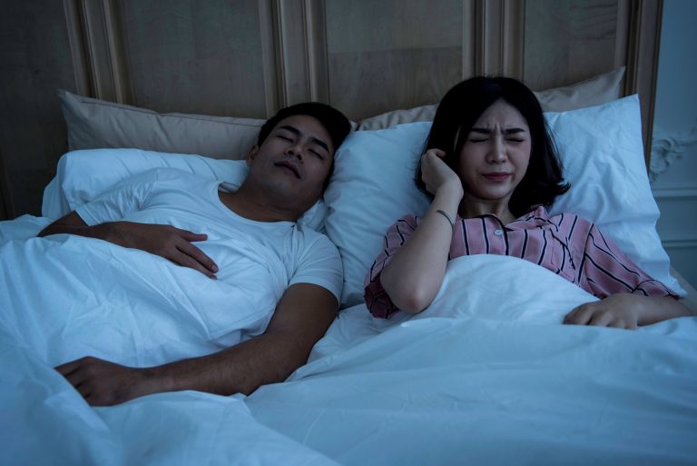 A woman covering her ears in discomfort while lying next to a man snoring, depicting the impact of sleep deprivation on couples